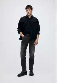 Ung mand stående iført en sort skjortejakke, sort polo-shirt, falmede sorte jeans, sort bælte og sorte sneakers mod en ensfarvet baggrund.