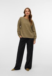 Olijfgroene sweatshirt met decoratieve parelaccenten, relaxte pasvorm en lange mouwen, gecombineerd met zwarte wijd uitlopende broeken en puntige beige schoenen.