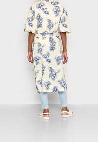 Kukkakuosiset keltaiset kimono-tyyliset viitat sinisillä kukilla ja lehdillä, vyötetty vyötäröltä, yhdistettynä vaaleansinisiin farkkuihin ja sandaaleihin.