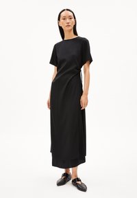 Schwarzes Maxikleid aus glattem Stoff, mit kurzen Ärmeln, Rundhalsausschnitt, seitlicher Bindedetail und geschichtetem Saum. Kombiniert mit flachen schwarzen Schuhen.