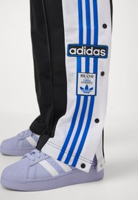 Svart och vit Adidas träningsbyxor med blå ränder och logotyper, i kombination med ljuslila Adidas sneakers som har vita ränder.