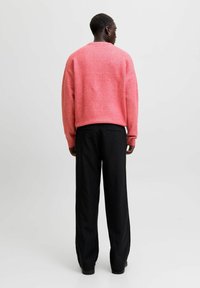 Persoon die met de rug naar de camera staat, gekleed in een losse roze trui en zwarte streepjesbroek tegen een effen lichte achtergrond.
