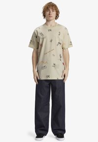 Camiseta beige con diseños gráficos en negro y rojo, combinada con pantalones de mezclilla oscuros de corte holgado y zapatillas negras. Conjunto de ropa casual.