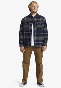 Camisa de franela a cuadros en azul marino con rayas beige y grises, dos bolsillos en el pecho y botones de bronce. Combinada con pantalones marrones y zapatillas negras.
