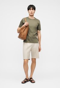 Fiatal férfi olívazöld pólóban, bézs shortban, sötétbarna szandálban, nagy barna bőr válltáskával a vállán, fehér háttér előtt áll.