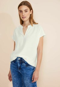 Giovane donna con capelli castano chiaro lisci, che indossa una blusa bianca a maniche corte e jeans in denim blu, su uno sfondo beige.