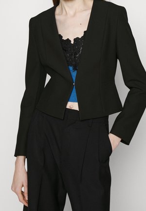 Kvinde iført en tætsiddende sort blazer over en blå top med sort blondekant og sorte bukser med høj talje, med den ene hånd i lommen.