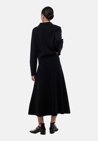 Un pull noir ajusté et une longue jupe fluide s'associent dans un tissu tricoté doux. La jupe présente un ourlet large et le modèle porte des chaussures noires brillantes.