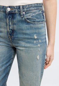 Μπλε τζιν παντελόνι με φθαρμένες λεπτομέρειες στον μηρό, μεταλλικό κουμπί, εμπρόσθιες τσέπες και μέρος του χεριού ενός ατόμου ορατό στο πλάι.