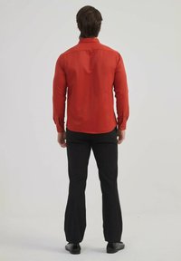 Chemise rouge à manches longues, texture lisse et ourlet arrondi, associée à un pantalon noir évasé et des chaussures sombres, vue de dos.