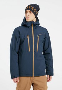 Chaqueta impermeable de color azul marino con cremalleras y acentos en tono beige, que cuenta con una capucha y múltiples bolsillos delanteros. Combinada con un gorro a juego.