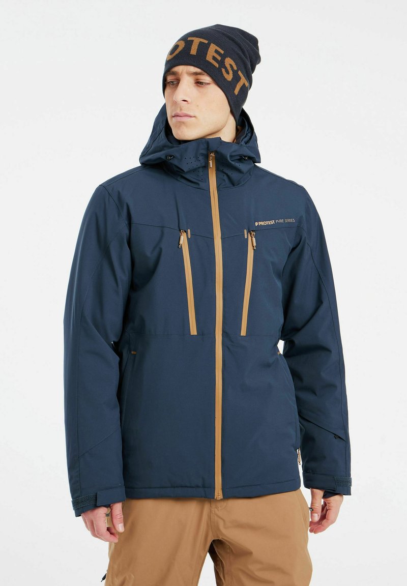 Chaqueta impermeable de color azul marino con cremalleras y acentos en tono beige, que cuenta con una capucha y múltiples bolsillos delanteros. Combinada con un gorro a juego.