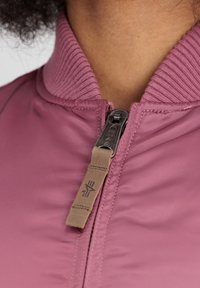 Veste bomber violet clair avec col côtelé et fermeture éclair. Dispose d'un onglet sur la tirette de la fermeture éclair et d'un tissu lisse, légèrement brillant.