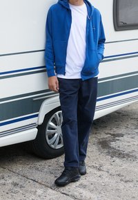 Sudadera azul con cremallera sobre una camiseta blanca, pantalones azul marino y zapatillas negras. El fondo presenta un vehículo blanco con rayas azules y grises.