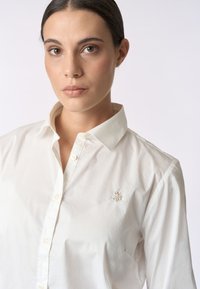 Camicia bianca con bottoni e colletto classico, realizzata in cotone morbido, con un piccolo logo ricamato sul lato sinistro del petto.