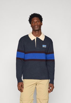 Σκούρος πουλόβερ με μπλε και μπορντό οριζόντιες ρίγες, μπεζ γιακά και λογότυπο στο στήθος. Πλεκτή υφή, χαλαρή εφαρμογή.