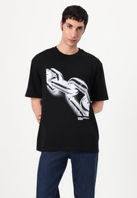 Juodas medvilninis marškinėlis su dideliu baltu grafikų vaizdu, pavaizduojančiu susijungusias grandines priekyje, o apačioje užrašyta "KARL LAGERFELD JEANS".