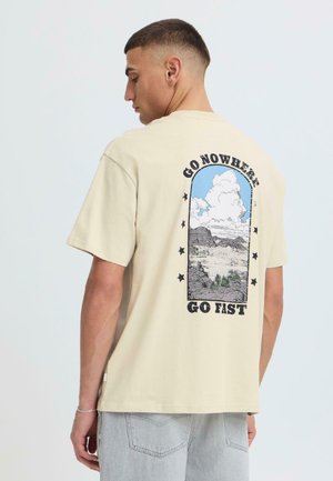 Jeune homme aux cheveux courts porte un t-shirt beige avec un paysage désertique, des nuages et le texte "GO NOWHERE GO FAST" au dos, associé à un jean clair.