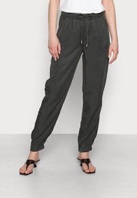 Pantalon gris foncé, léger et fuselé, avec une taille élastique et un cordon de serrage. Comprend des poches latérales et une texture douce. Porté avec des sandales marron.