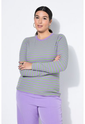 Kobieta z ciemnymi włosami związanymi do tyłu, ubrana w zielono-fioletową pasiastą bluzkę z długim rękawem, lawendowe spodnie, złote kolczyki koła i pierścionki, stojąca z założonymi rękami.