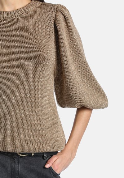 Pull en maille marron avec manches bouffantes aux coudes porté avec un pantalon noir, main posée de manière décontractée dans la poche, sur fond clair.
