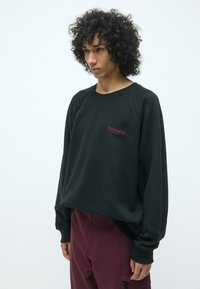 Sudadera negra con diseño de raglán, puños de canalé y un pequeño logo bordado en burdeos. Tejido suave con un corte relajado.