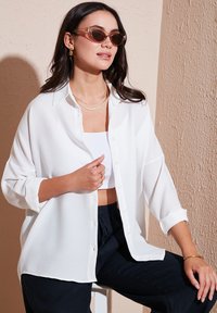 White, textured shirt with buttons and roll-up sleeves, paired with a white cropped top and dark pants. Accessories include sunglasses and layered necklaces.