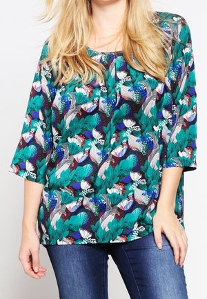 Kvinde iført en løs, langærmet bluse med et farverigt blomster- og bladmotiv i grøn, blå, hvid og lilla, kombineret med blå jeans.