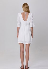 Robe blanche courte avec un dos nu, des manches évasées et des points texturés, comportant des accents en dentelle le long de l'encolure et de l'ourlet. Liens au dos.