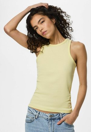 Jonge vrouw met krullend haar, een mouwloos geel topje en een blauwe spijkerbroek, één hand in haar zak en de andere raakt haar hoofd aan.