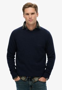 Granatowy sweter dzianinowy z okrągłym dekoltem, noszony na zielonej koszuli w kratę, w zestawie z niebieskimi dżinsami. Swobodny i warstwowy wygląd.