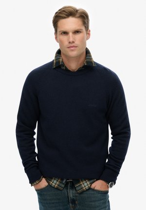 Granatowy sweter dzianinowy z okrągłym dekoltem, noszony na zielonej koszuli w kratę, w zestawie z niebieskimi dżinsami. Swobodny i warstwowy wygląd.