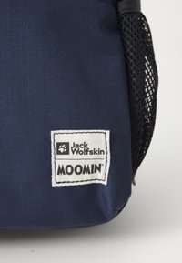 Mörkblå ryggsäck med ett strukturerat tyg, som har en vit etikett med "Jack Wolfskin" och "MOOMIN" logotyper samt en svart nätficka på sidan.