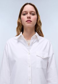 Camicia bianca con bottoni realizzata in cotone, dotata di una singola tasca sul petto, colletto appuntito e tessuto leggero con una consistenza liscia.