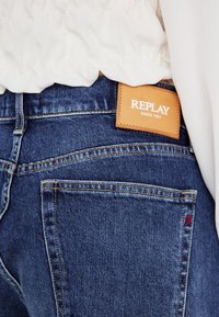 Džínové kalhoty v tmavě modré barvě s hladkou strukturou, s výrazným žlutým prošíváním, nápadnou hnědou visačkou s logem "REPLAY" a červeným akcentem.