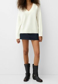 Biały sweter z dekoltem w serek, z prążkowanymi mankietami i krawędzią, zestawiony z ciemną dżinsową mini spódnicą oraz czarnymi kozakami do kolan z srebrnymi ozdobami.