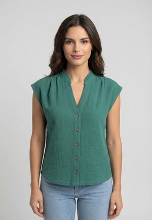 Mujer con cabello largo y oscuro, vestida con una blusa verde sin mangas con botones y jeans azul claro, de pie frente a un fondo gris liso.