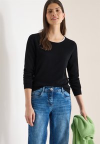 Femme portant un chemisier noir à manches longues et un jean bleu, tenant un pull vert plié dans sa main droite sur un fond neutre.