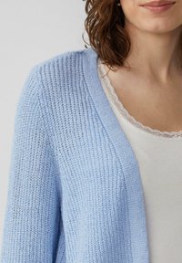 Lichtblauwe gebreide cardigan met een gestructureerd, opengewerkt ontwerp, voorzien van een brede kraag. Gekocht over een witte ribbeltop met kantafwerking.