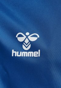 Una tela azul presenta un logo blanco con un diseño estilizado de abeja sobre el nombre de la marca "hummel" en letras claras. Textura suave.