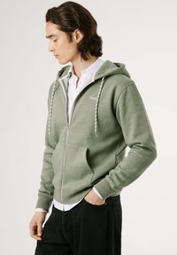 Sudadera verde con cremallera, hecha de tela suave, con detalles de cordón, bolsillos laterales y un logo en el pecho. Lleva puesta sobre una camiseta blanca.