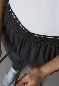 Schwarze Laufshorts mit einem elastischen Bund, der ein weißes Muster mit dem Text "RUNNING" aufweist, sowie verstellbaren Kordelzügen. Graue Sportschuhe sind sichtbar.