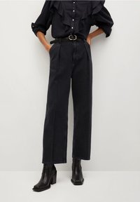 Blusa negra con volantes y botones, combinada con pantalones de mezclilla oscuros de talle alto y pernera ancha, y botas de tacón negro con punta. 