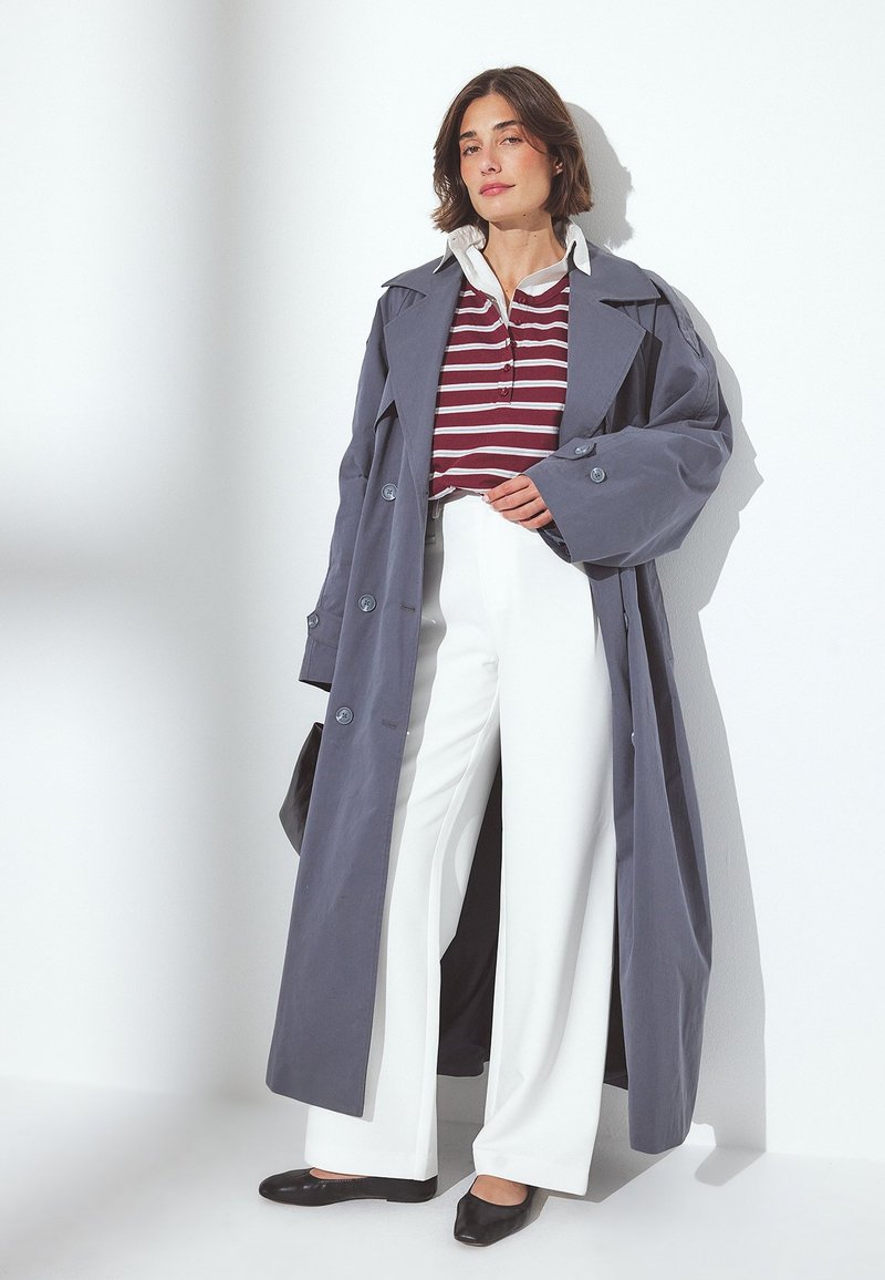 Długi szary trencz, białe spodnie w szerokie nogawki, bordowa bluzka w paski oraz czarne płaskie buty. Gładki materiał z guzikami i kołnierzykiem.