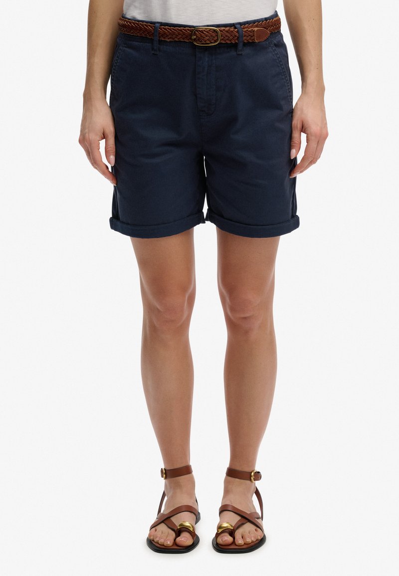 Mujer con pantalones cortos azul marino con dobladillo enrollado, cinturón trenzado marrón y sandalias marrones de tiras, de pie contra un fondo blanco.