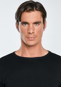 Jaunas vyras su trumpais rudo plaukų ir mėlynomis akimis, dėvintis juodą apvalios apykaklės marškinėlį, žiūrintis tiesiai į priekį prieš tuščią baltą foną.