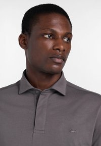 Szara polo z miękkiego materiału, z zapięciem na guziki u kołnierza oraz haftowanym logo psa z lewej strony. Gładkie, dopasowane kroje.