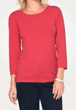 Femme portant un haut rouge uni à manches trois-quarts et un pantalon sombre, avec un bracelet en argent au poignet droit, debout devant un fond blanc.