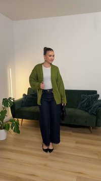 Grønn oversized cardigan, grå topp, mørke vide jeans, svarte hæler, poserer i et lyst rom med planter og en grønn sofa.