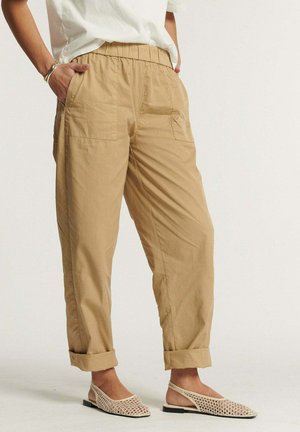 Personne portant un pantalon beige retroussé avec poches avant, une chemise blanche, un bracelet argenté et des ballerines slingback tissées beige, debout devant un fond uni.